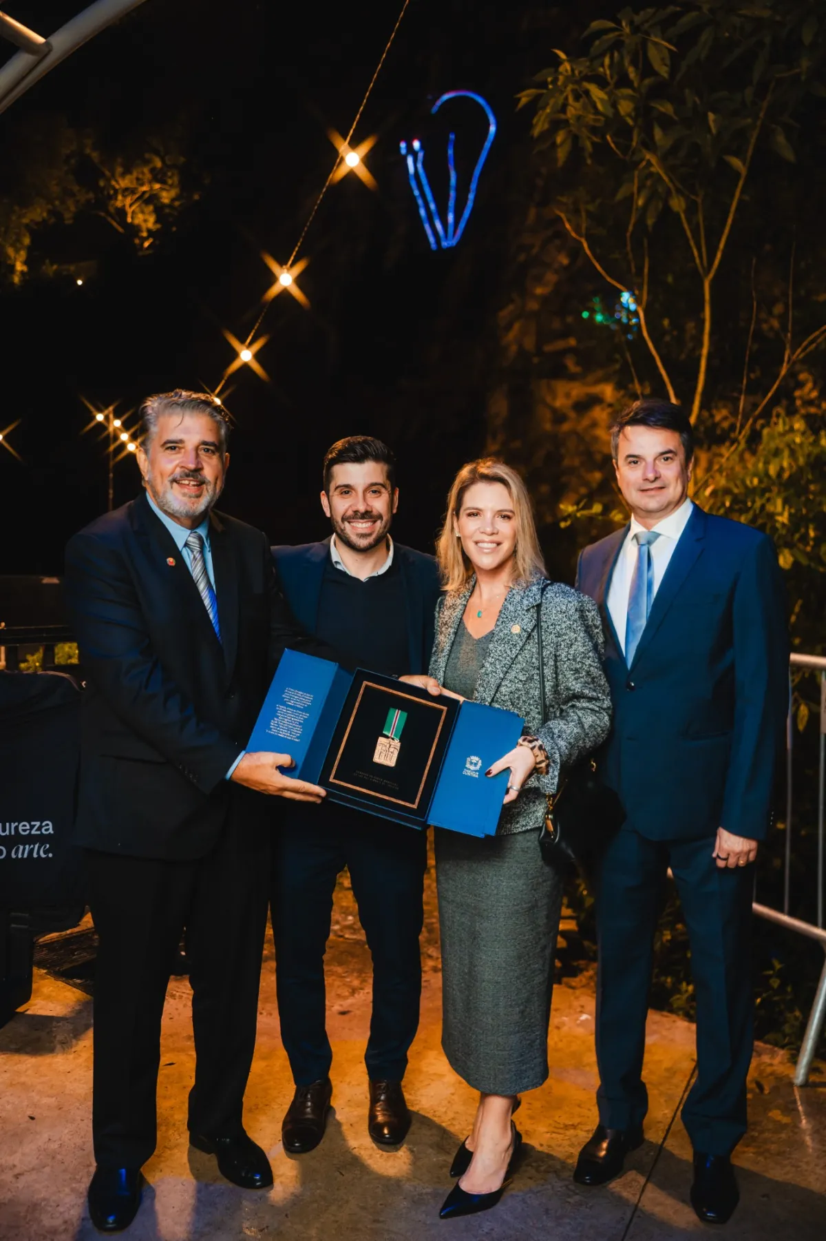 Délio, Caio Canabrava, Tatiana Reichmann e o marido segurando a comenda, ao ar livre, sob um varal de luzes