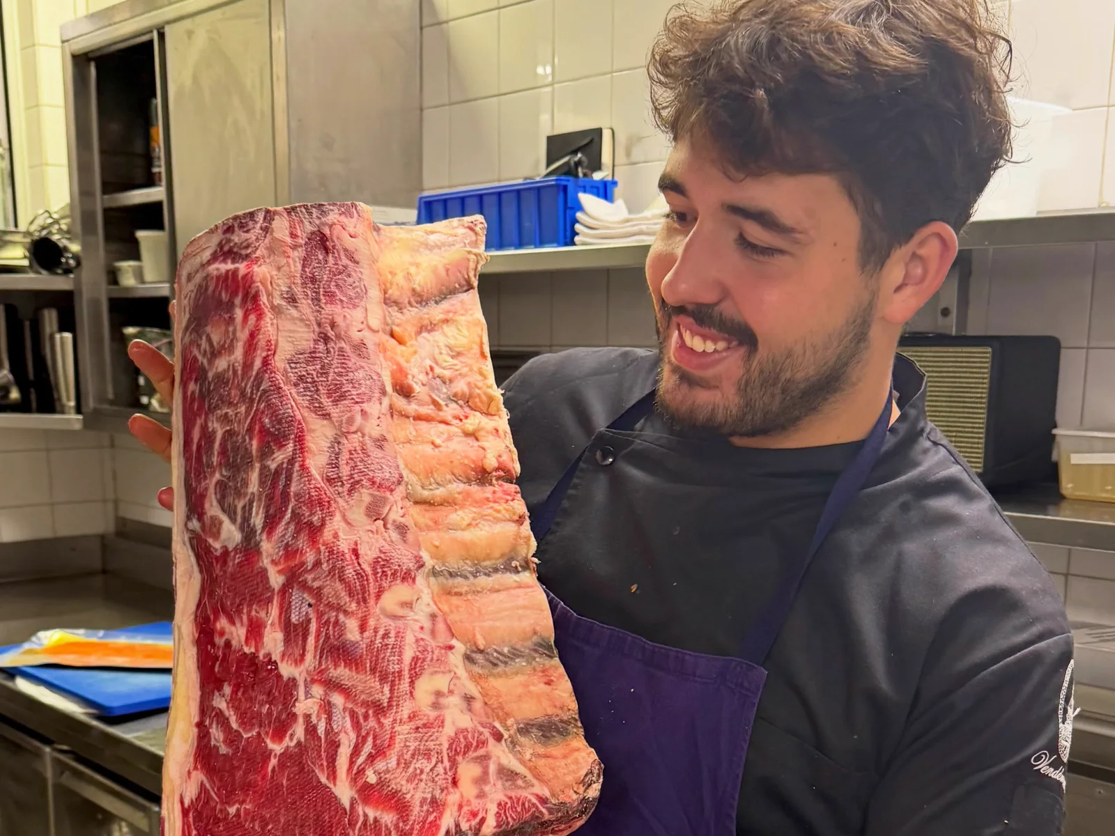 Pedro Canabrava sorri segurando uma peça inteira de costela na cozinha da brasserie, em Paris